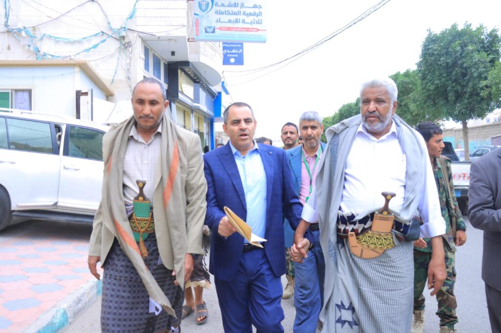 1001114393 - جامعة 21 سبتمبر Group of men walking down a street in a procession; two men wear traditional Yemeni clothing with daggers at their waists, while a man in a blue suit holds a folder in the center.