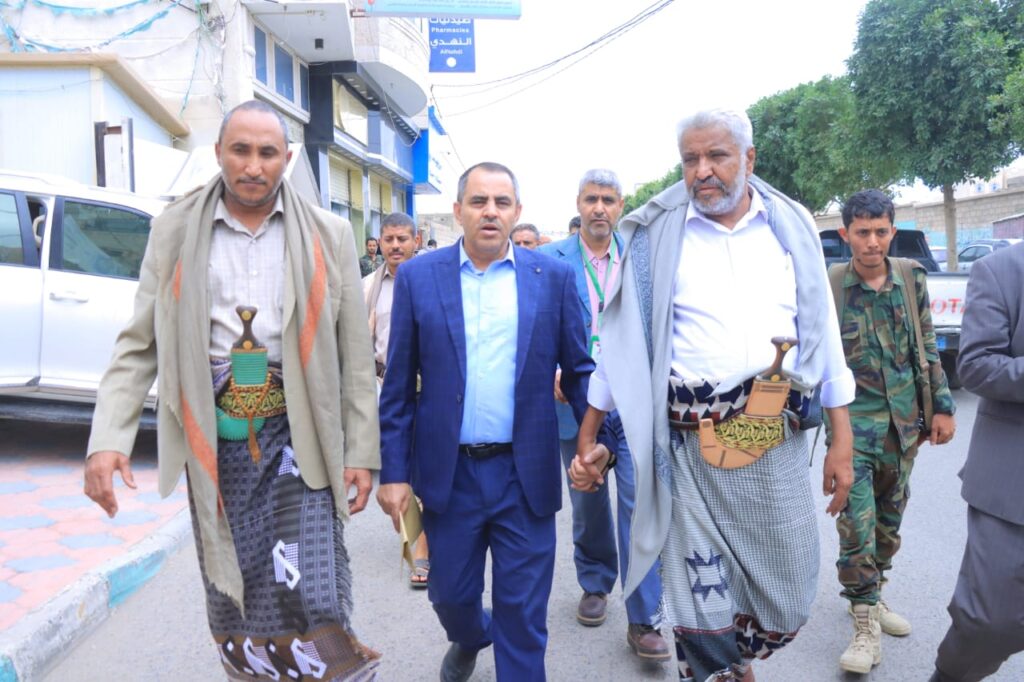 1001114396 - جامعة 21 سبتمبر Group of men walking on a city street; two in traditional belt daggers flank a man in a blue suit.