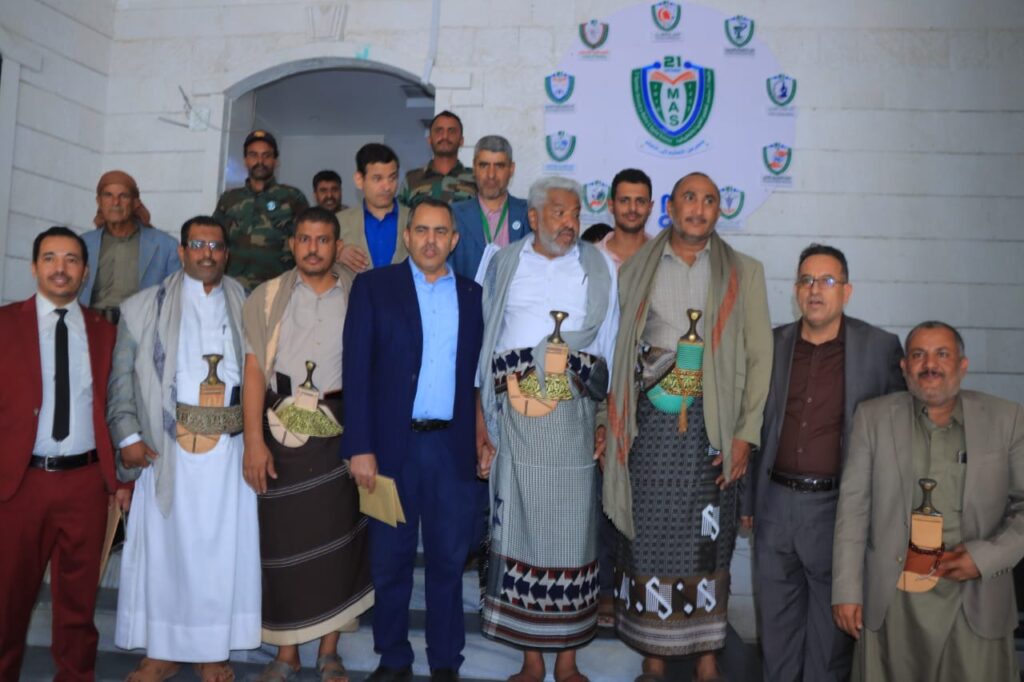 1001114399 - جامعة 21 سبتمبر Group of men in traditional Yemeni dress and suits pose for a photo at an event, with ceremonial daggers at their waists.