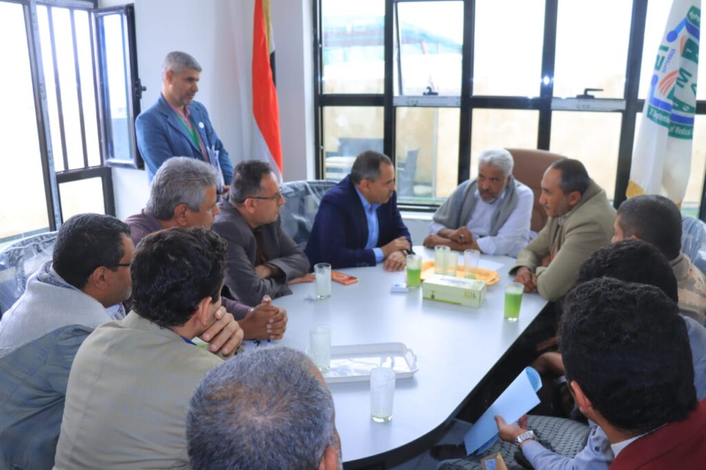 1001114405 - جامعة 21 سبتمبر Group of men seated around a white conference table in a bright room, having a discussion with drinks and a tissue box on the table.