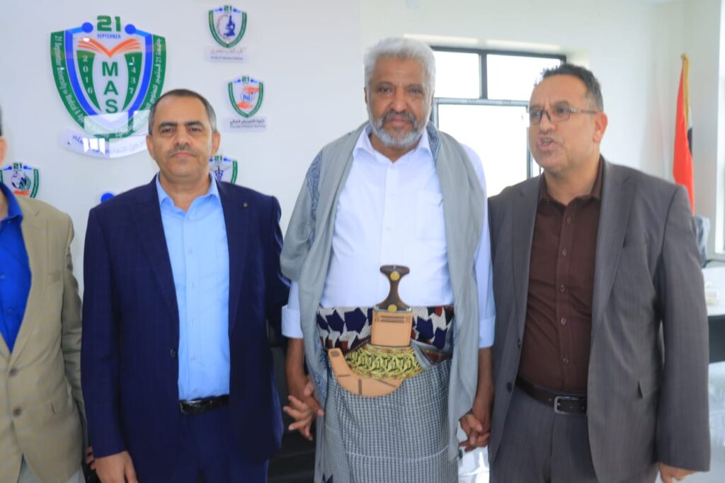 1001114412 - جامعة 21 سبتمبر Group of three men posing at a formal event; center man wears traditional Yemeni clothing and a dagger belt.