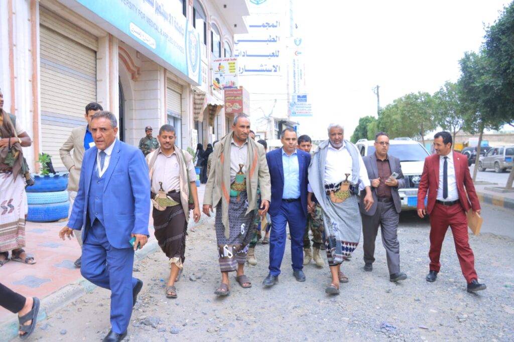 1001114417 - جامعة 21 سبتمبر Group of men in suits and traditional clothing walk along a dusty city street with storefronts and parked cars behind them.
