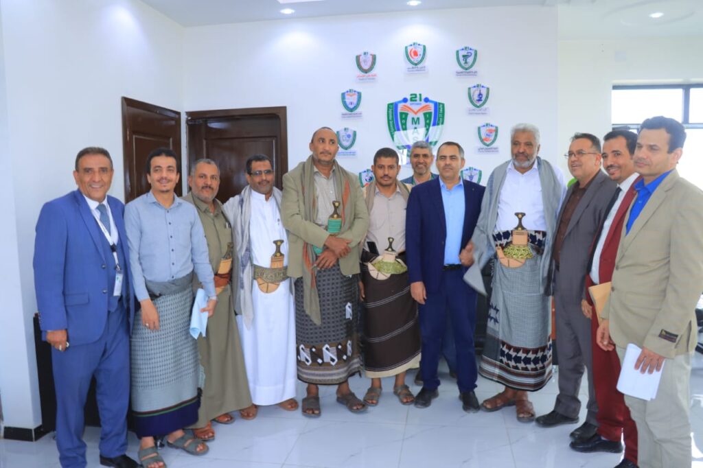 1001119861 - جامعة 21 سبتمبر Twelve men in traditional and business attire pose for a group photo in a bright room with crest logos on the wall.