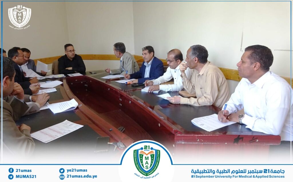 Group of professionals seated around a long conference table, reviewing papers in a meeting room with a university emblem visible on the overlay border.