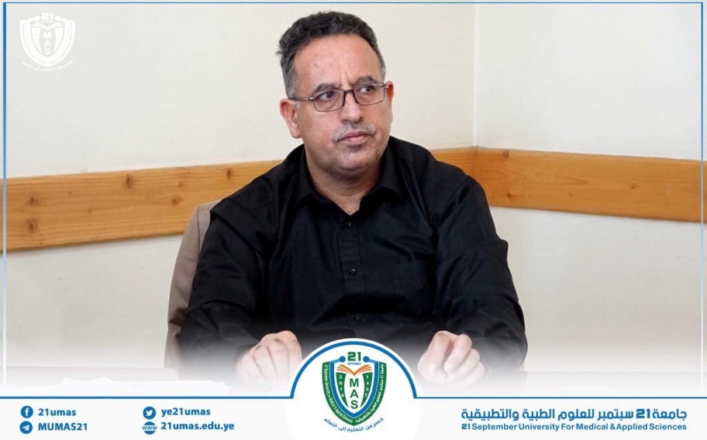 Man in a black shirt wearing glasses sits at a table in a meeting room, thoughtful expression, with a university-branded banner and seal at the bottom edge.