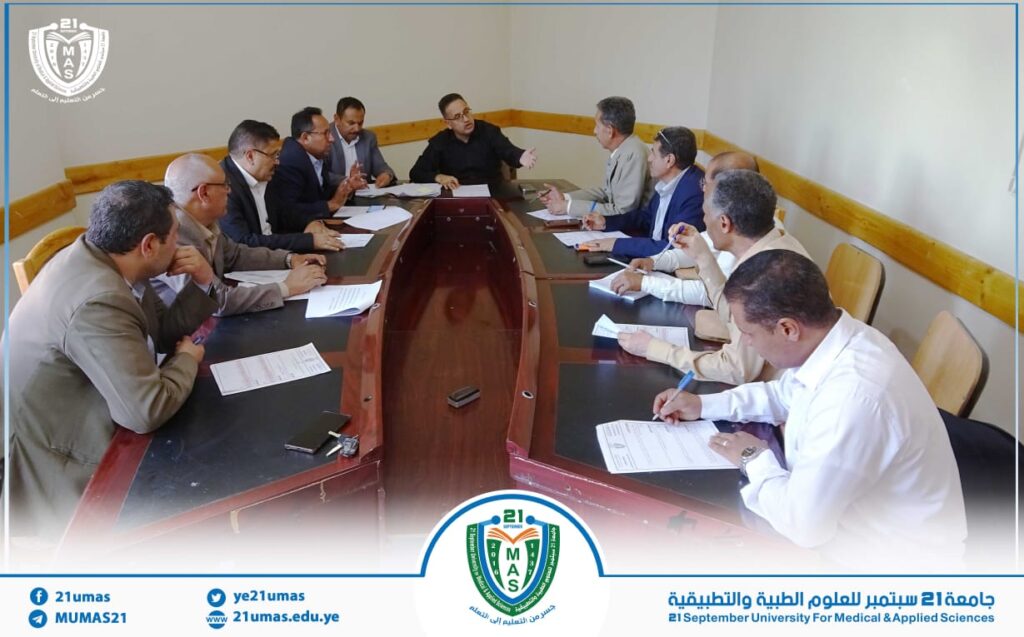 Group of men in a formal meeting around a U-shaped conference table, reviewing documents, with a university banner at the bottom.