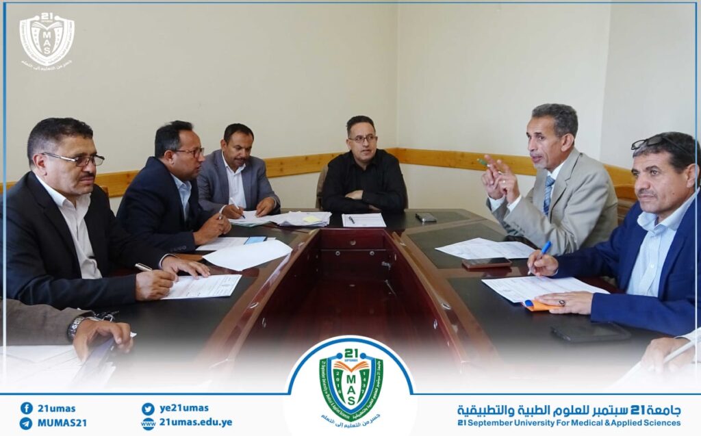 Group of professionals seated around a conference table, discussing with papers and pens in a formal meeting at a university setting. A crest logo and blue border are visible in the frame.