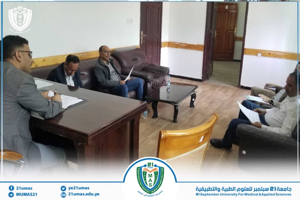 Group of men in a meeting room around a large desk and a sofa, reviewing documents; university branding visible on the banner at the bottom and a watermark in the corner.