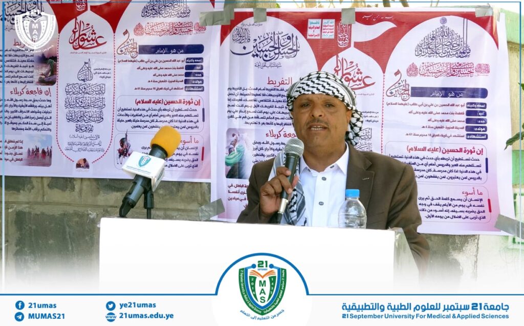 Man in a brown blazer and keffiyeh speaks into a microphone at a podium, with Arabic posters and a water bottle behind him at a public event.