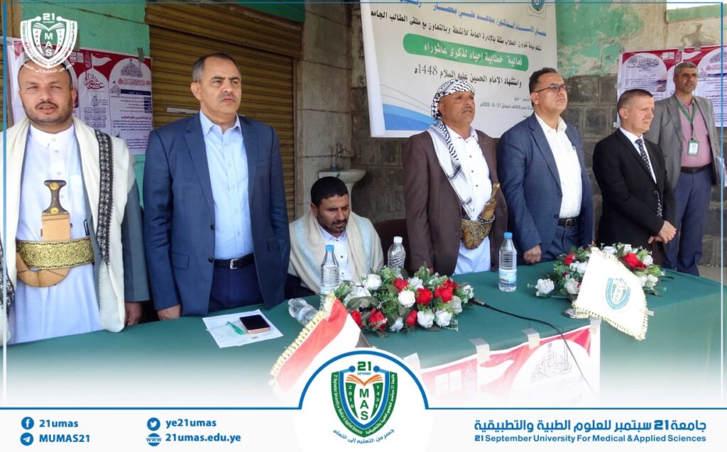 Group of men in suits and traditional dress standing behind a decorated table at a university ceremony, with a banner and the 21mas logo in the background.