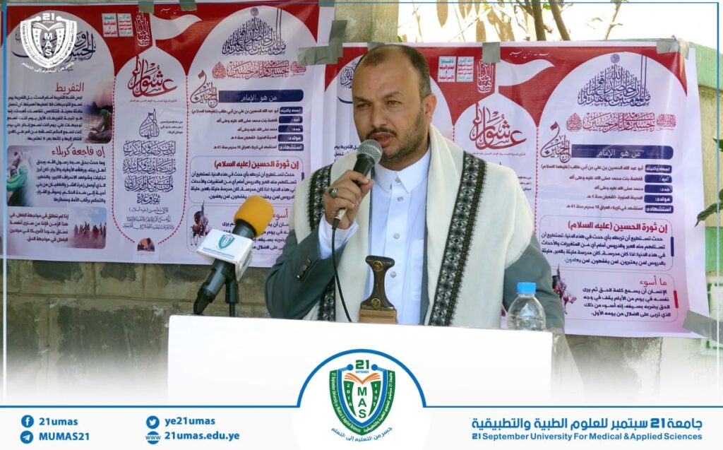 Man speaking into a microphone at a podium during a university event, with Arabic posters and banners in the background.