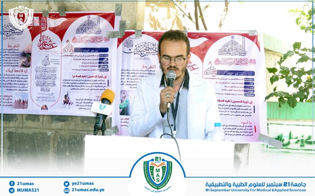Male speaker in a white coat delivering a talk at a podium with two microphones, posters in Arabic behind him, and a university banner at the bottom.
