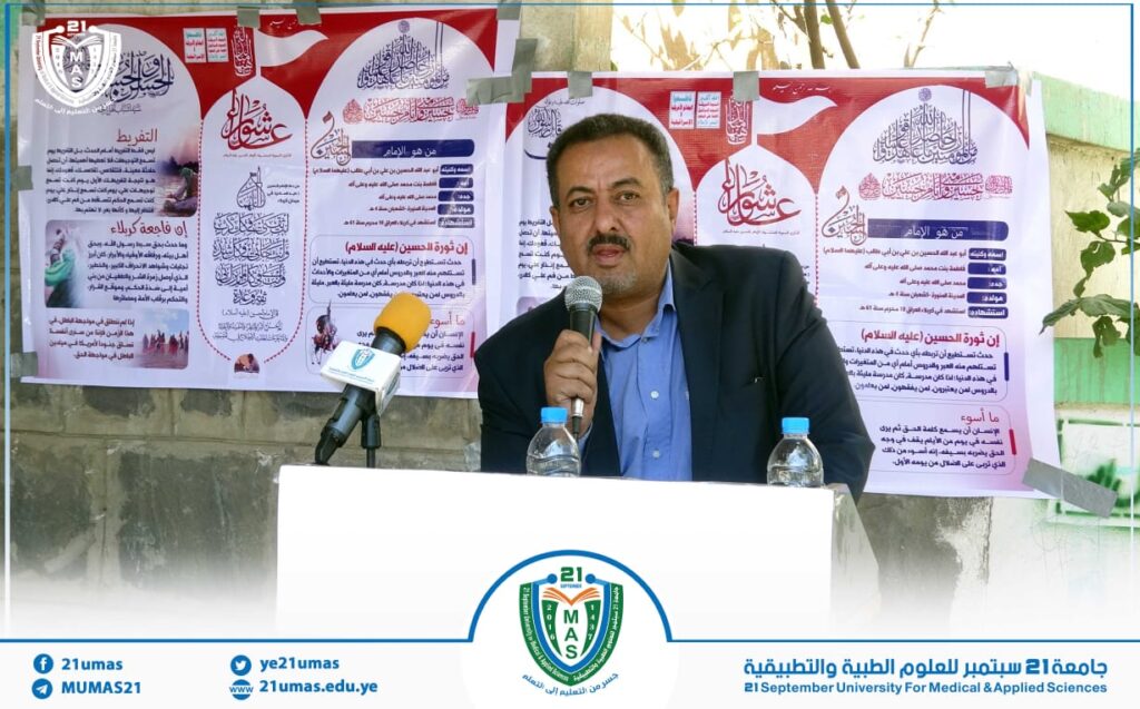 Man in a suit and blue shirt speaks into a handheld microphone at a white podium, with large Arabic posters behind him.