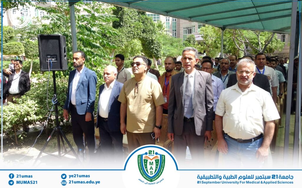 Group of formally dressed men standing under a canopy at a university event, facing forward near a microphone and speaker.