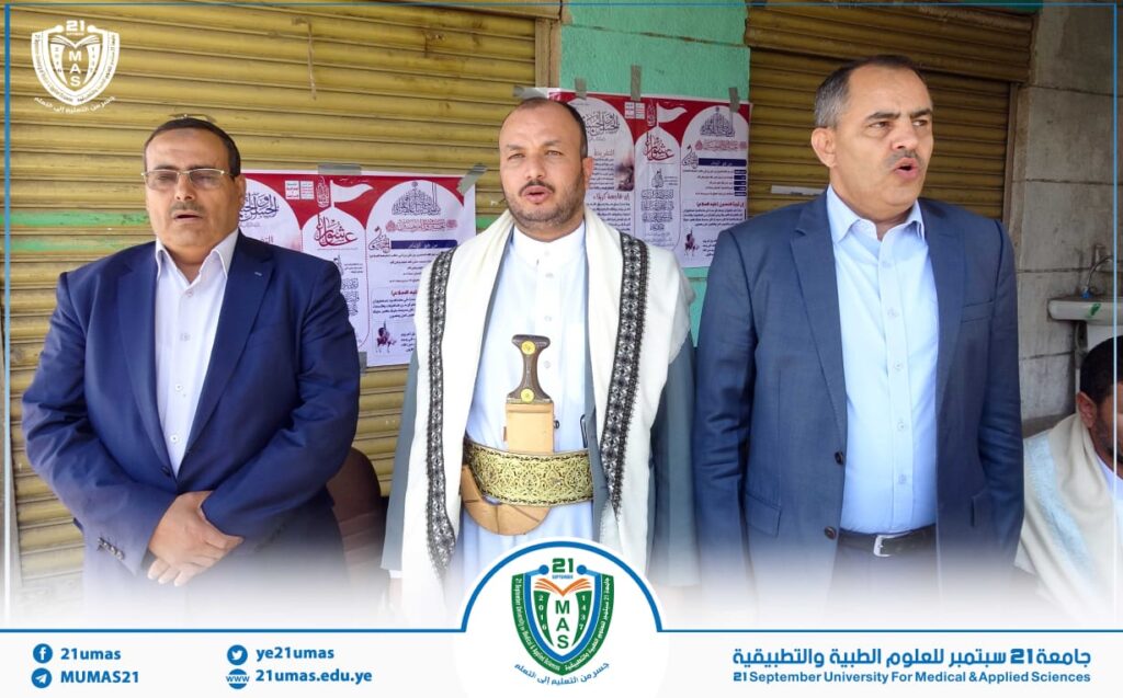 Three men in suits stand side by side at a storefront event; the central man wears traditional Yemeni attire with a ceremonial sword belt, posters on the wall behind them.