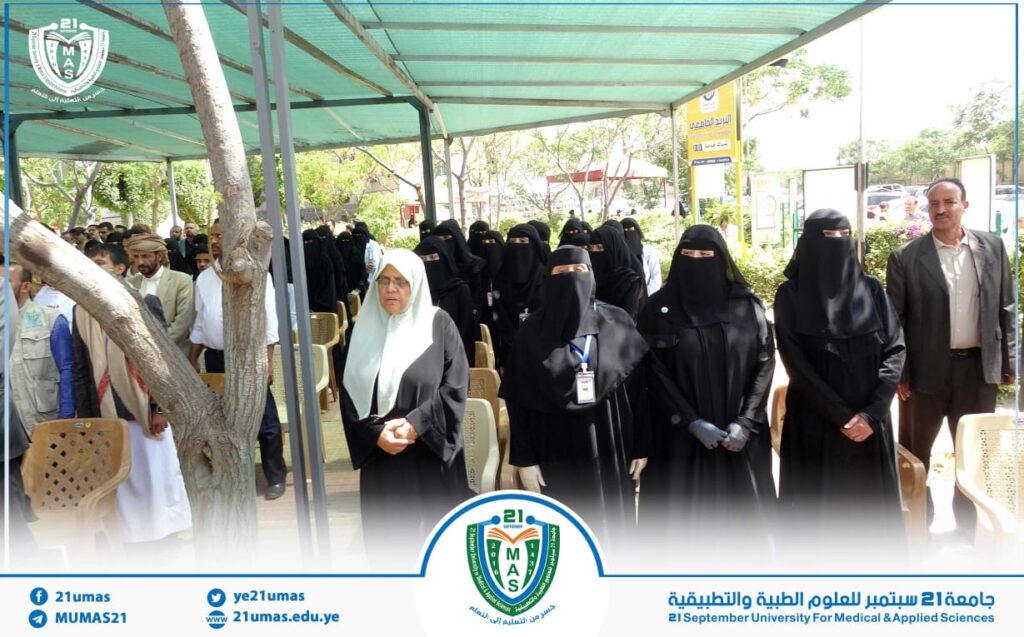 Group of women in black abayas and niqabs standing in rows at an outdoor university event under a green canopy, with an elderly woman in a white hijab at the front.