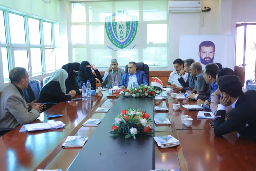 Group of professionals seated around a long conference table as one man speaks, with a floral centerpiece and a flag on the table in a meeting room behind them.