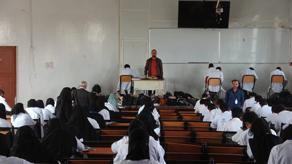 Lecture hall scene: a man in a brown jacket and red shirt stands at a desk at the front as students in white coats sit at benches facing forward in a large classroom.