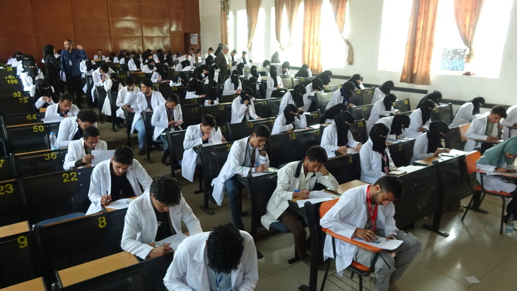 Large classroom filled with students in white coats taking an exam, many wearing black hijabs, seated in numbered rows across a bright room.
