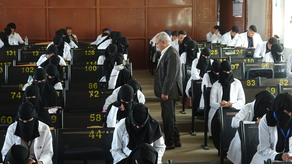 Older man in a gray suit walks through a crowded exam hall as students in white coats and black hijabs work at numbered desks