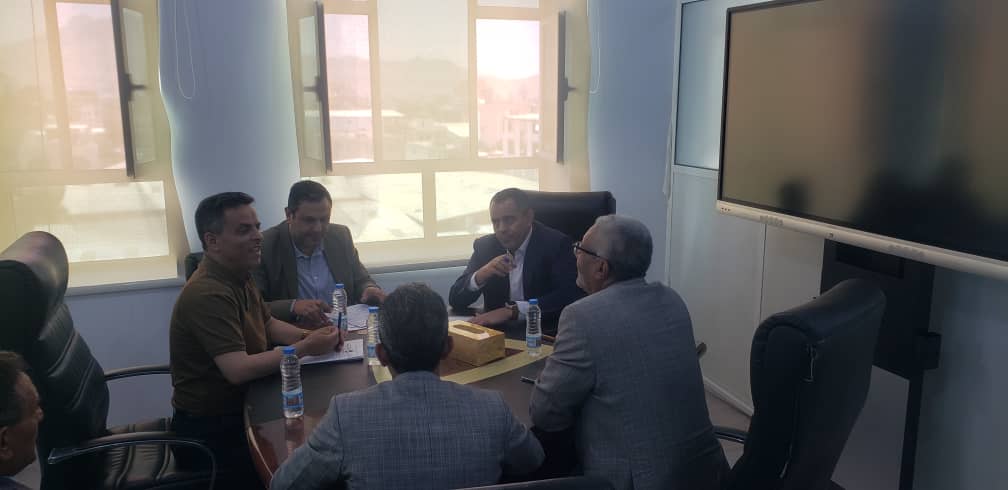Six professionals sit around a wooden conference table in a bright meeting room, with water bottles and a wall-mounted display screen nearby.