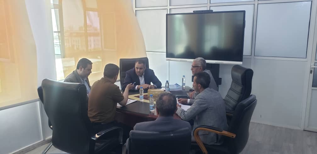 Six men in a conference room seated around a dark wood table, discussing with papers and water bottles nearby, and a large screen on the wall behind them.