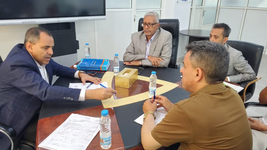 Four men in business attire sit around a conference table reviewing documents, with pens, water bottles, and a large screen in the background.