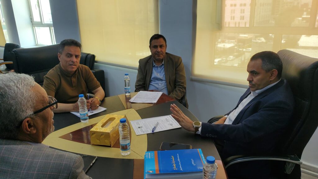 Group of four men in a business meeting around a conference table with papers and bottled water.