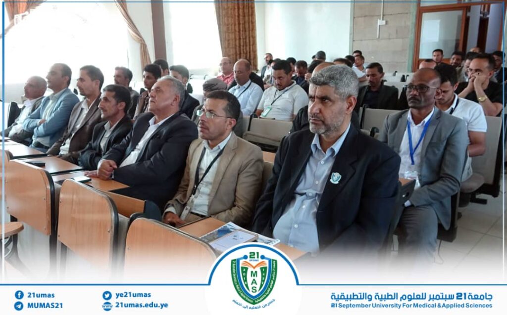 Group of professionally dressed men seated in a lecture hall attending a university conference; university crest visible at the front edge of the image.