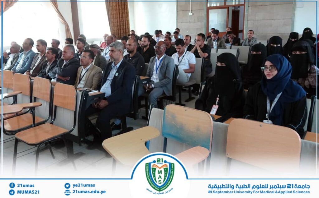 Audience seated in a university lecture hall for a conference or seminar; mix of men in suits and women in hijabs in the front rows, with a university logo and Arabic banner framing the bottom of the image