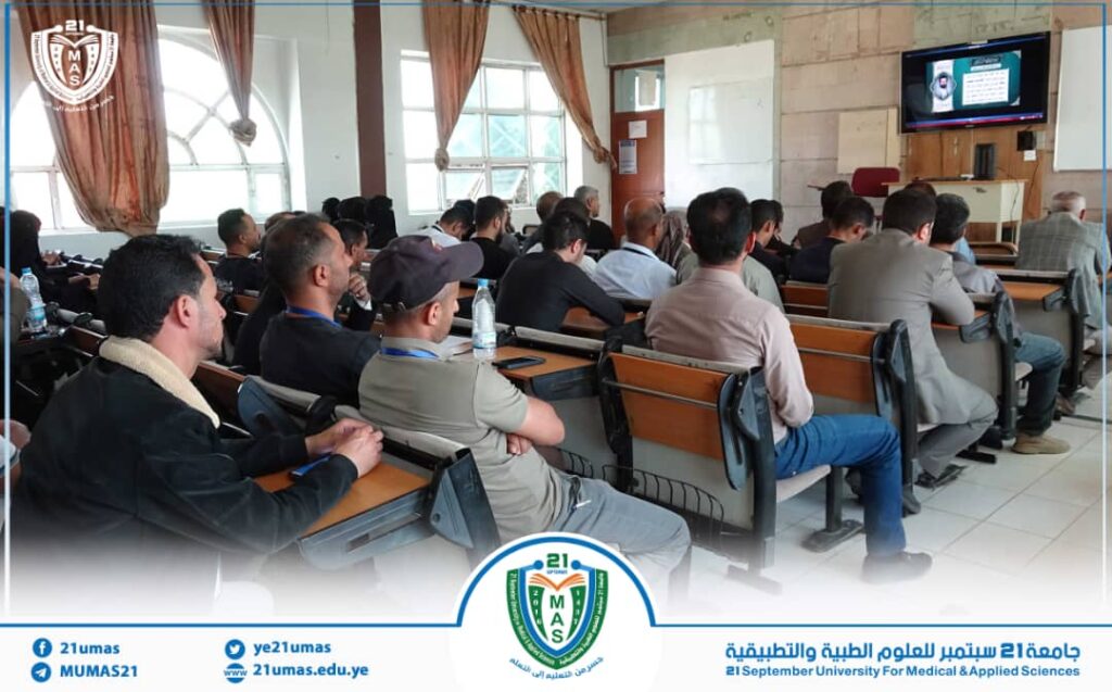 Students in a university lecture hall watching a presentation on a screen; visible logo and banner in the room.