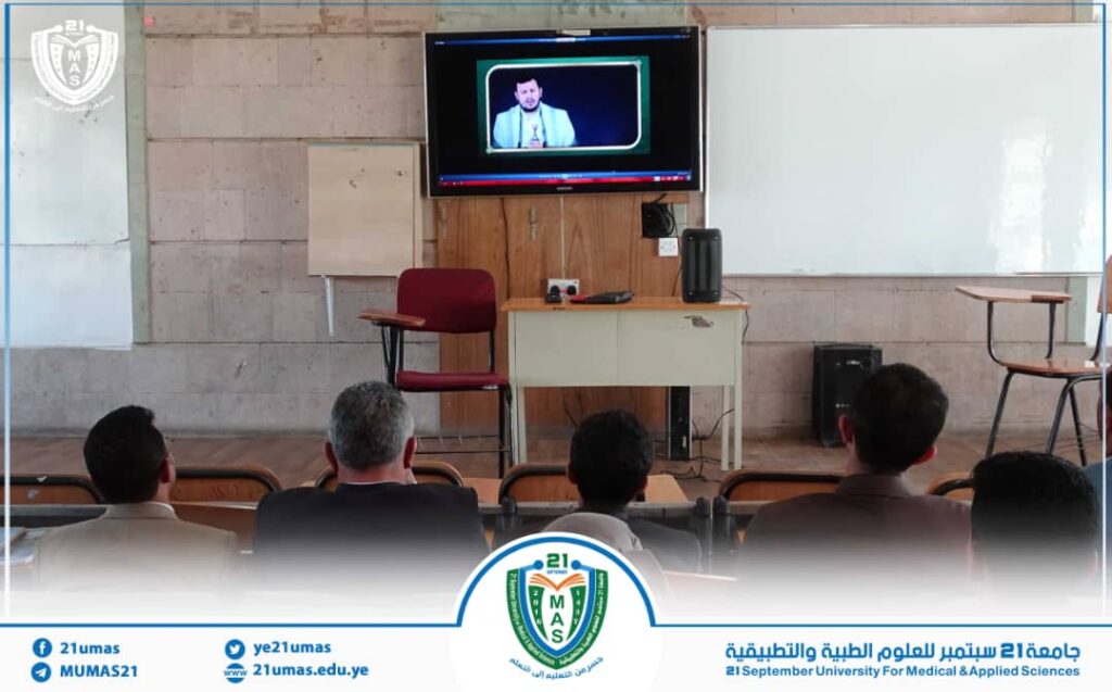 Lecture hall setup: a speaker appears on a mounted TV screen above a desk facing an audience of students.