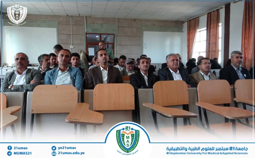 Group of men in suits seated in a university lecture hall, facing the front, with a crest/logo visible at the bottom of the frame.