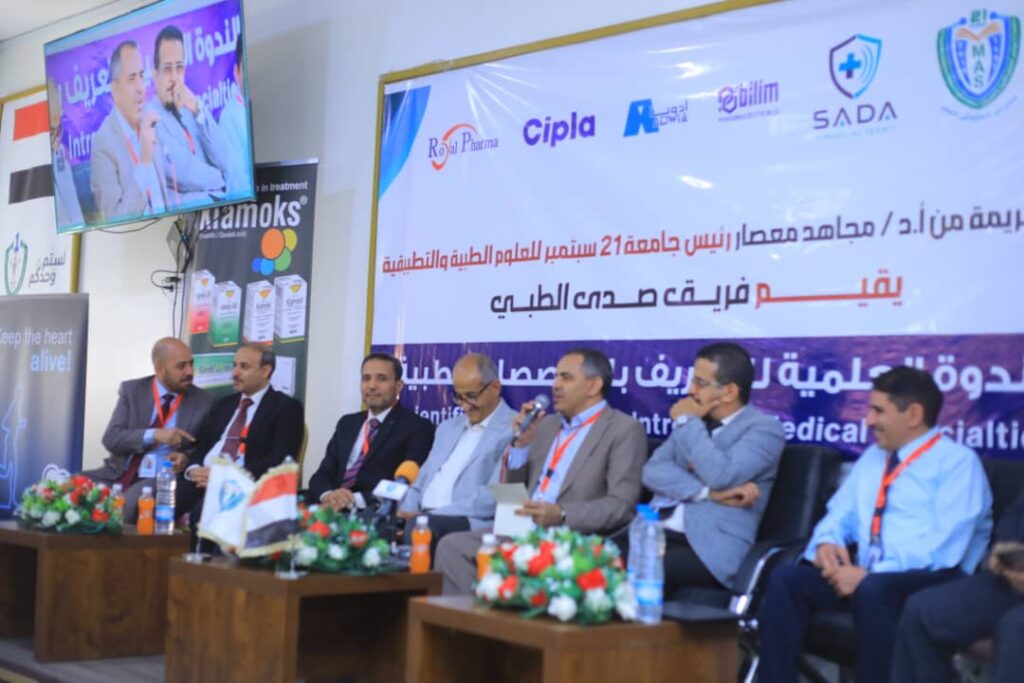 Panel of six men in suits seated on stage at a medical conference, with a banner containing Arabic text behind them.