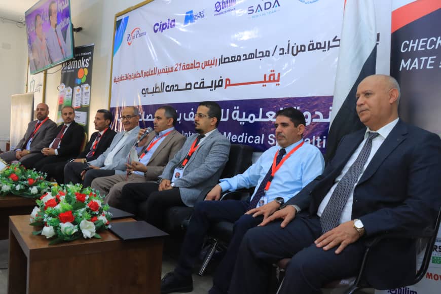 Panel of men in suits seated on a stage during a medical conference, with a large Arabic banner behind them and floral arrangements in front.