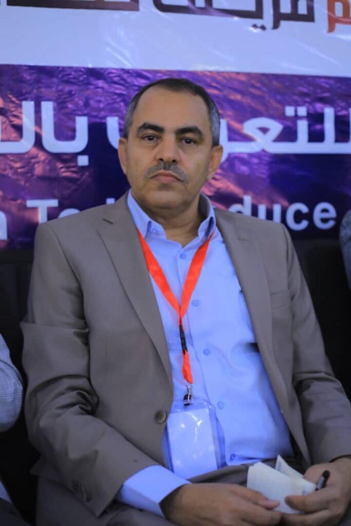 Middle-aged man in a gray suit with a blue shirt, wearing an orange lanyard, seated at a conference panel.