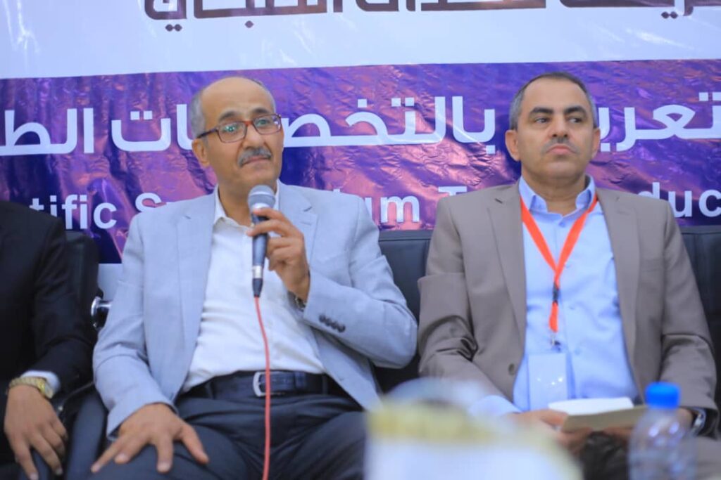 Two men in suits seated on a panel stage; one holding a microphone, speaking, with a purple banner in the background.