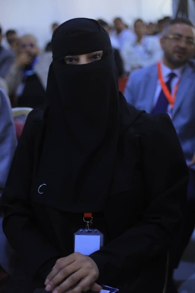 Person in a black niqab with a lanyard badge seated at a conference or event.