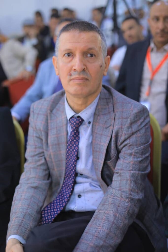 Middle-aged man in a gray plaid blazer and blue shirt, wearing a patterned tie, seated at a conference with blurred attendees in the background.