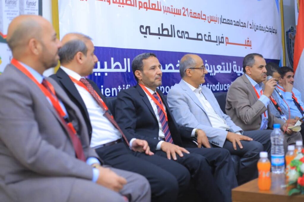 Group of suited men seated in a panel on a stage at a conference, with a blue and white banner behind them.