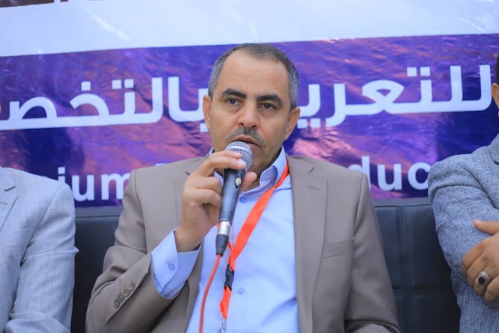 Middle-aged man in a beige suit speaking into a microphone at a panel, with a purple banner in Arabic behind him.