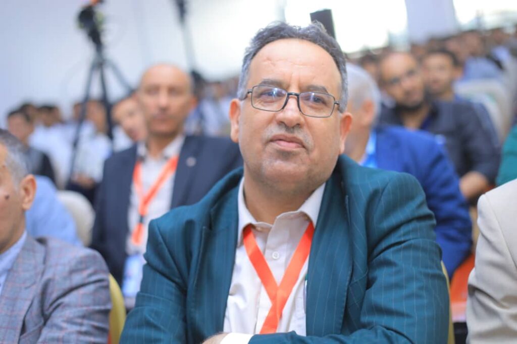 Man wearing glasses in a teal pinstripe suit with an orange lanyard at a conference setting.