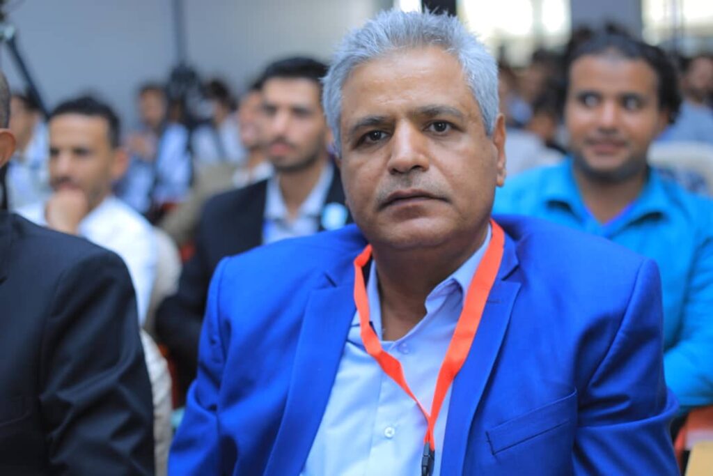 Man in a blue suit and light blue shirt wearing an orange lanyard sits at a conference, with blurred attendees in the background.