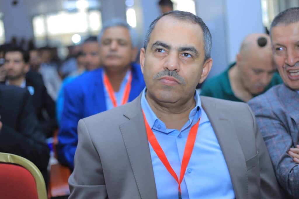 Middle-aged man in a gray suit and blue shirt wearing a red lanyard at a crowded conference discussant? event.