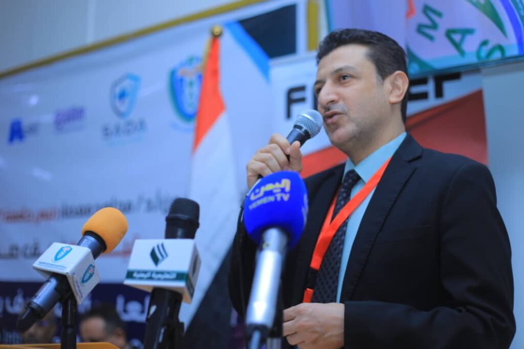 Man in a suit speaking into a microphone at a press conference, with multiple mic banners in front of him and a backdrop behind.