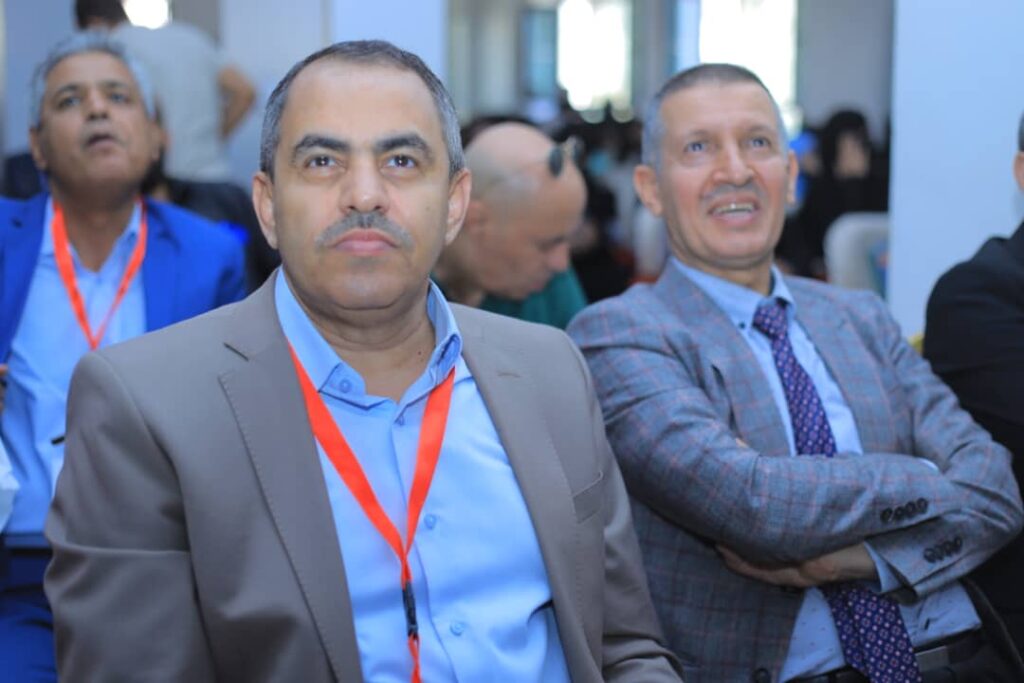 Two men in business attire seated at a conference; foreground man in a tan blazer with a blue shirt and red lanyard, beside a smiling man in a gray plaid jacket.