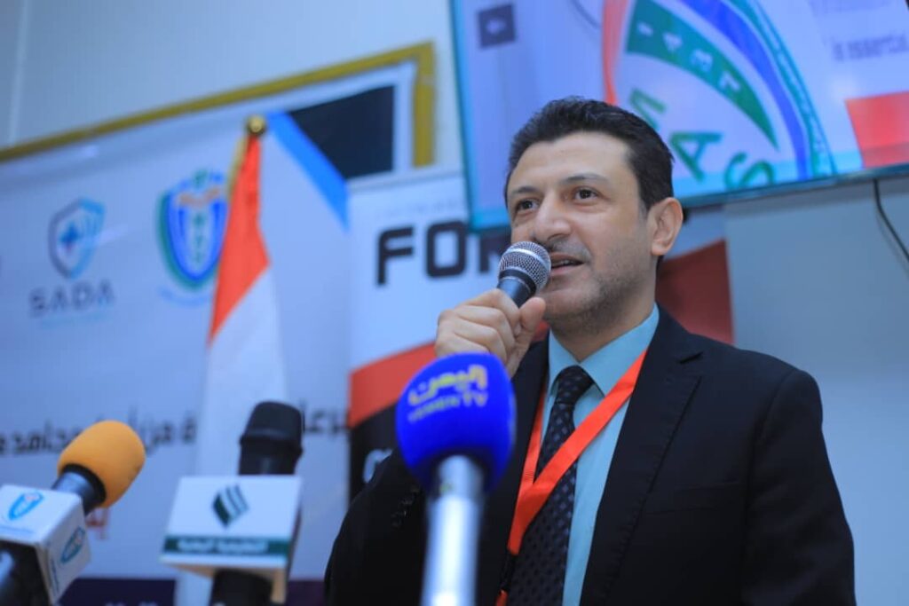 Man in a dark suit and blue shirt speaks into a handheld microphone at a press conference, with several microphones in front and banners in the background.