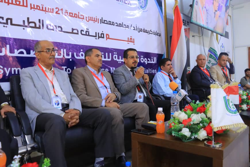Panel of six men in suits on a conference stage, speaking and listening, with microphones, a large Arabic banner, Iraqi flag, and a floral centerpiece on the table.