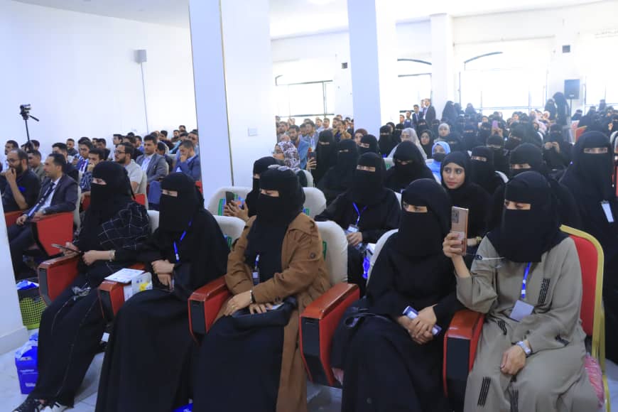 Audience seated in a conference hall; many women wearing niqab, some using phones, listening to a presentation.