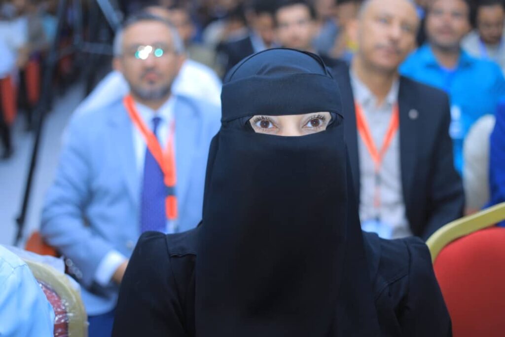 Woman wearing a black niqab with only her eyes visible, seated in a conference audience.
